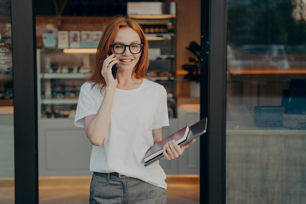 Smiling woman with laptop and notebook in hand enjoying talk on cellphone near coffee shop